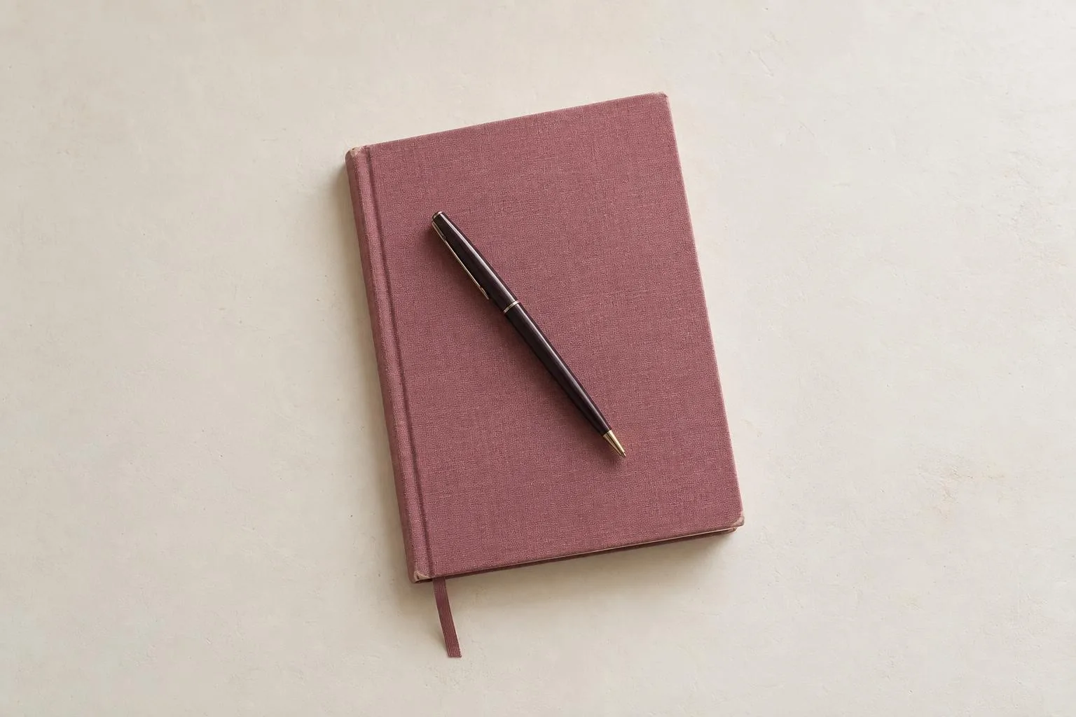 A closed cloth-bound notebook in a dusty rose binding with a slim dark pen resting across it.